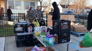 People sorting donated food items on outdoor tables near a building, with crates and various groceries visible.