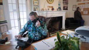 Person in colorful shirt on phone at desk; another person stands near bulletin board in room with fireplace and plants.