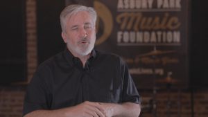 A man in a black shirt speaks in front of a sign that reads "Asbury Park Music Foundation.