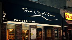 Blue awning of "Town & Surf Diner" with contact number. Brick building with illuminated windows and a pizza sign nearby.