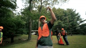 A construction worker in an orange vest and hard hat gestures upward in a wooded area.