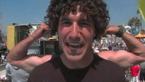 Person smiling in foreground with curly hair; another flexing their arms in the background at a crowded outdoor event.