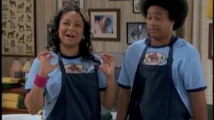 Two people wearing blue shirts and aprons stand in a room with framed pictures on the walls.