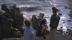 Three people stand on rocky cliffs by the ocean, facing the crashing waves.